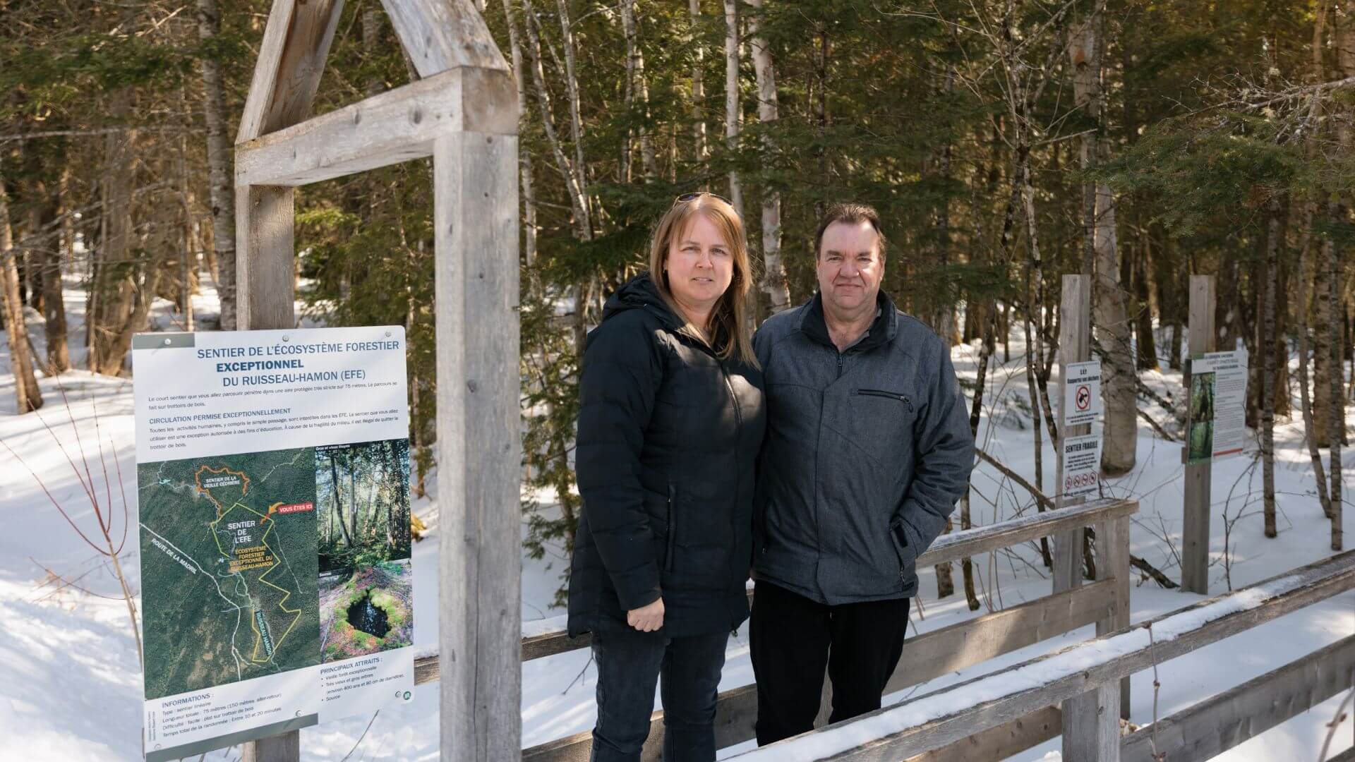 Deux personnes à l’entrée du sentier de la forêt ancienne du Ruisseau-Hamon, à Saint-Marcel, devant un panneau d’interprétation.